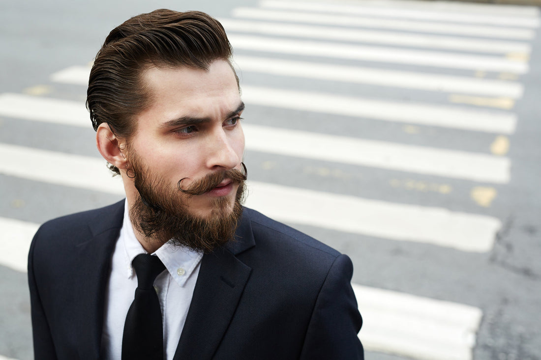 Bearded man in a dark suit standing at an outdoor crossing with a sharply shaped beard