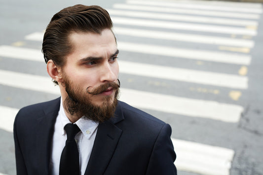 Bearded man in a dark suit standing at an outdoor crossing with a sharply shaped beard
