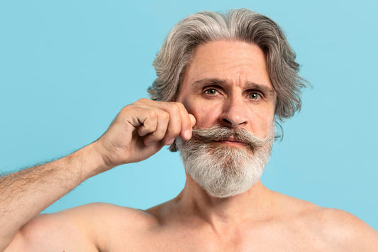 Older man with a full grey beard twirling the end of his mustache against a pale blue backdrop